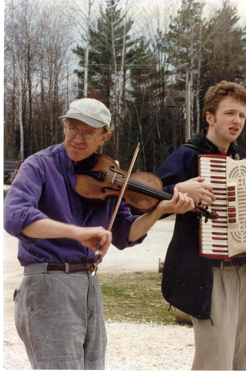 An man playing a fiddle outdoors with a young man holding an accordion next to him, in a wooded area.