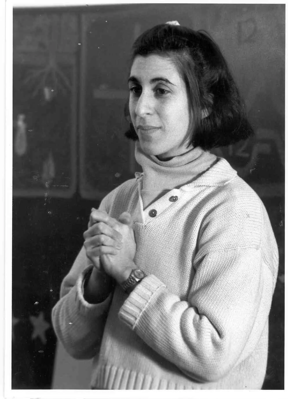 A woman with dark hair, wearing a light-colored sweater and turtleneck, clasping her hands together in front of her chest, looking contemplative.