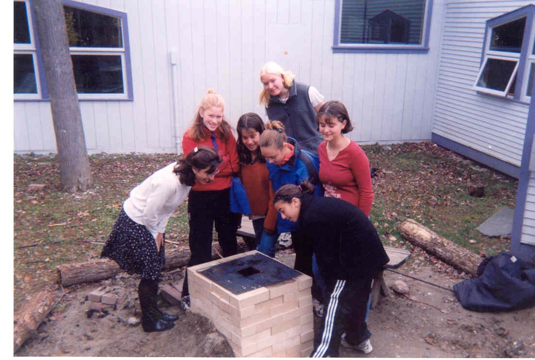 Group of children and an adult gathered around a small brick outdoor fire pit in a backyard.
