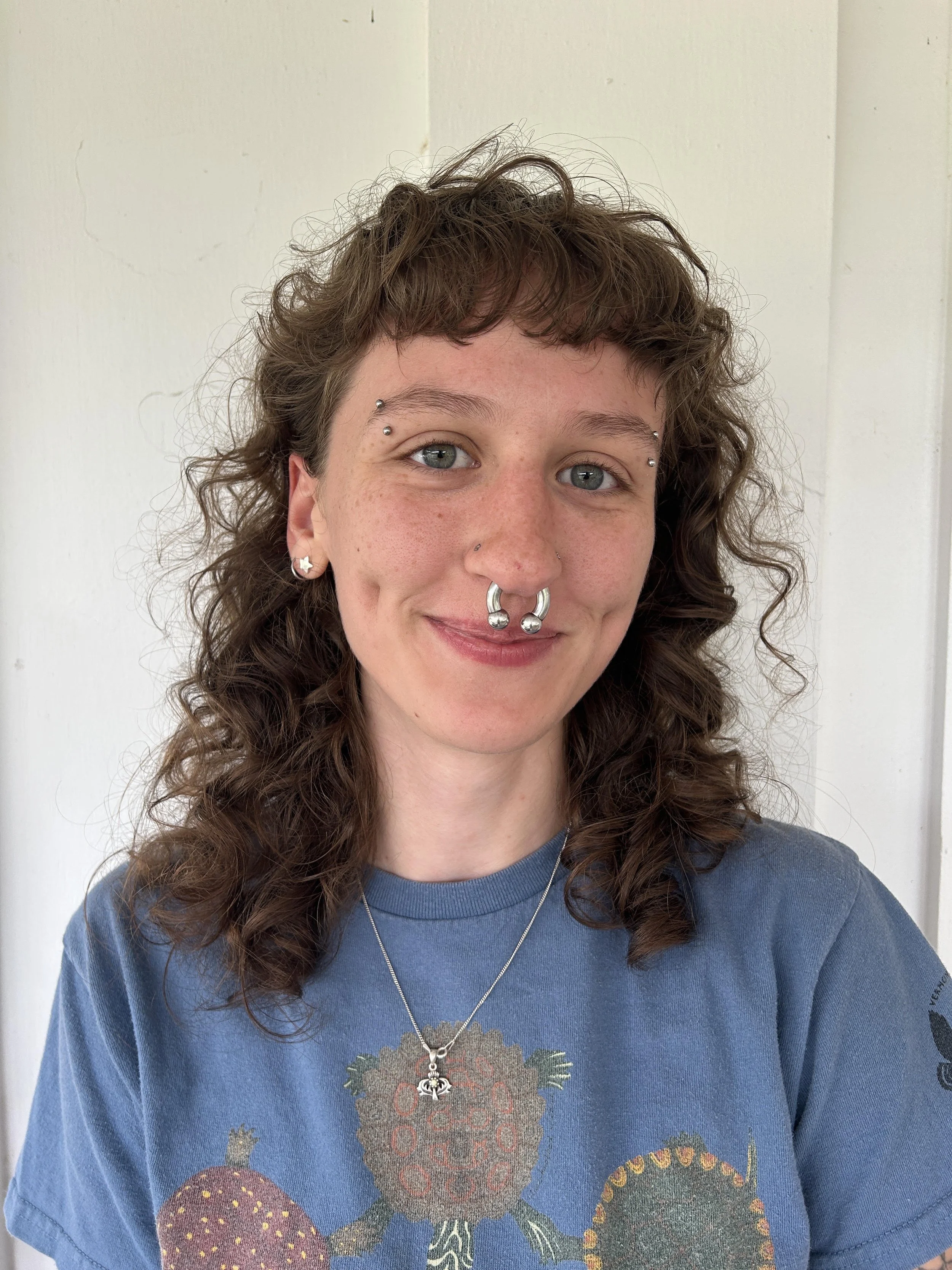 A young woman with curly brown hair and multiple facial piercings, wearing a blue t-shirt with a floral print, a necklace with a bee pendant, and small earrings, smiling in front of a plain white wall.