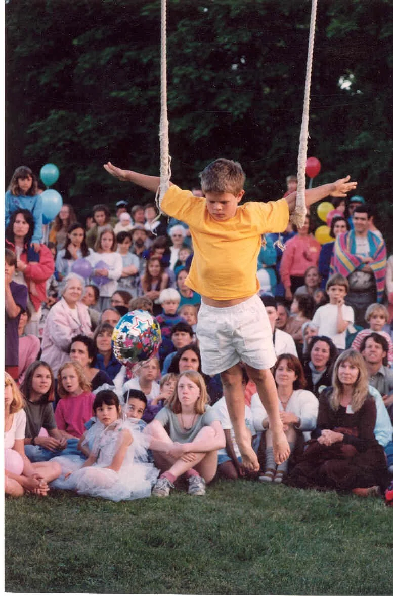 Young boy in orange shirt and white shorts swinging on a rope swing at an outdoor gathering, surrounded by a seated and standing crowd of children and adults, some holding balloons.