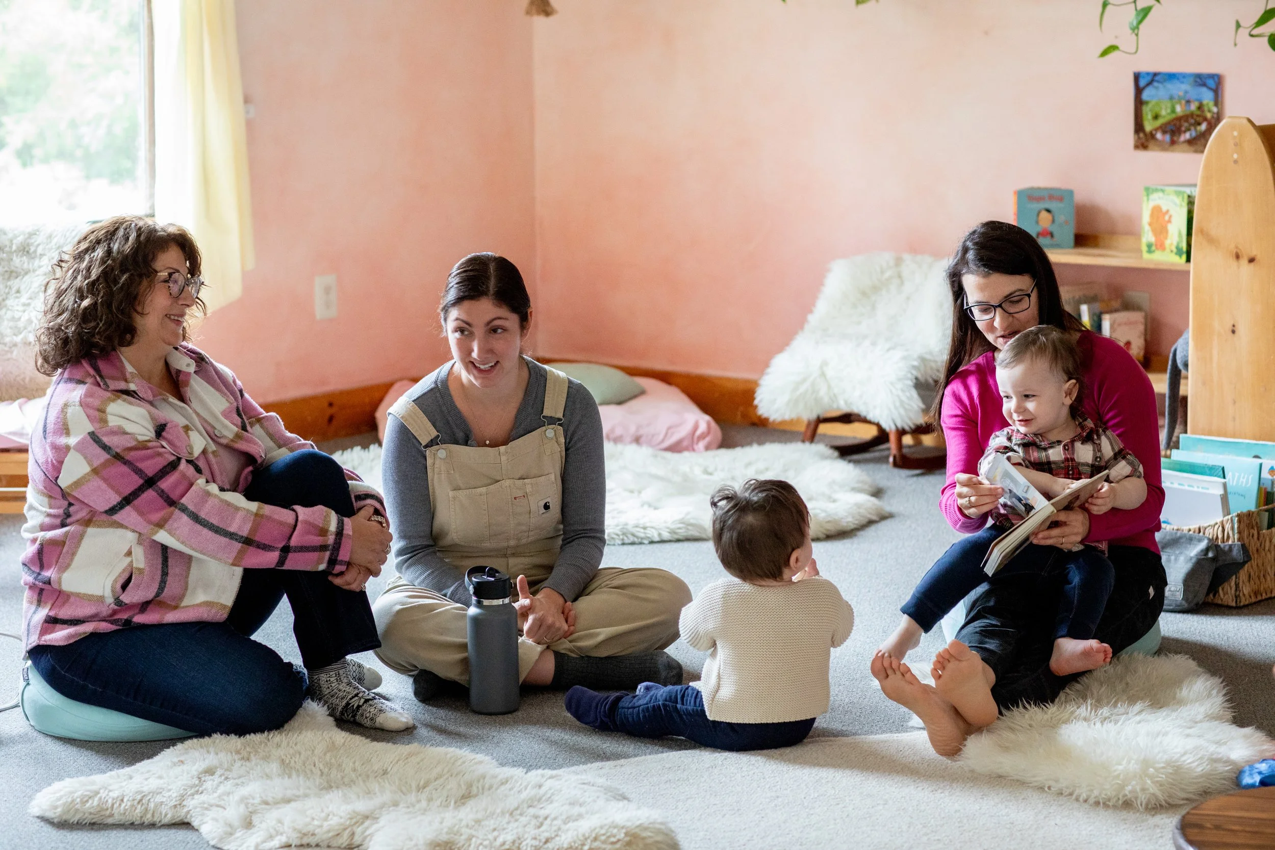 Three mothers and their babies laughing in a pink classroom with sheep's wool on the ground