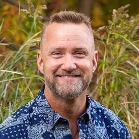 A smiling man with short hair and a beard, wearing a blue patterned shirt, standing outdoors with tall grasses and plants in the background.