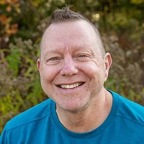 A smiling man outdoors with trees in the background, wearing a blue shirt.