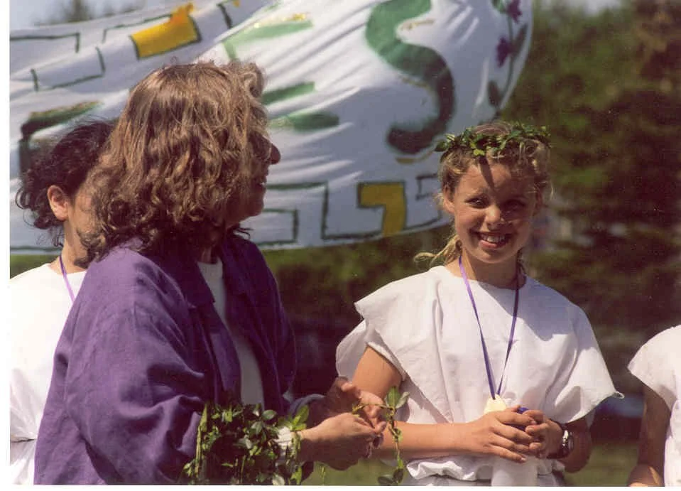 A young girl with a laurel of leaves on her head, smiling and holding a small branch, standing next to a woman with curly hair wearing a purple jacket, at an outdoor event with a banner in the background.