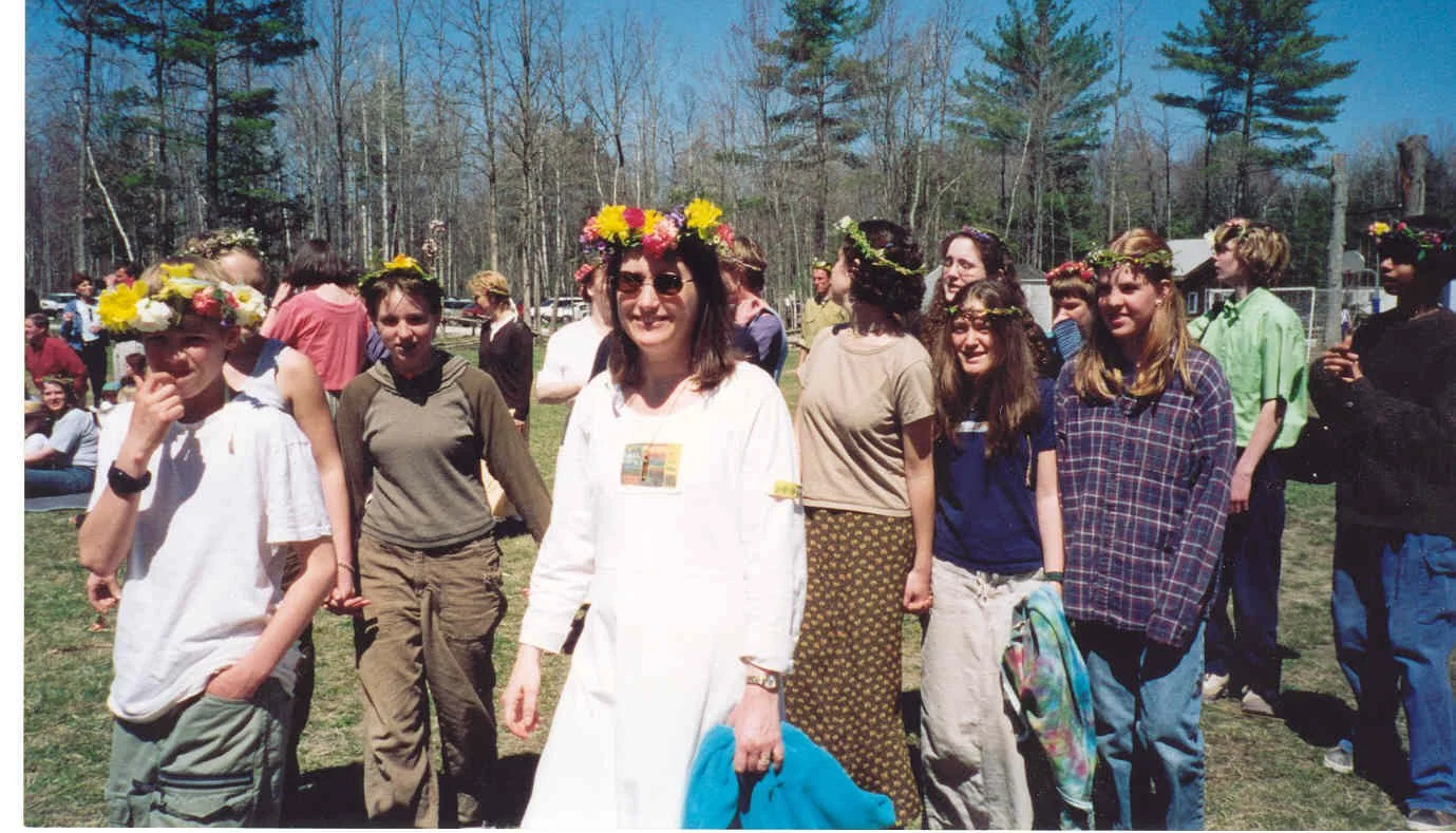 Group of young women wearing flower crowns outdoors on a sunny day, with trees and more people in the background.