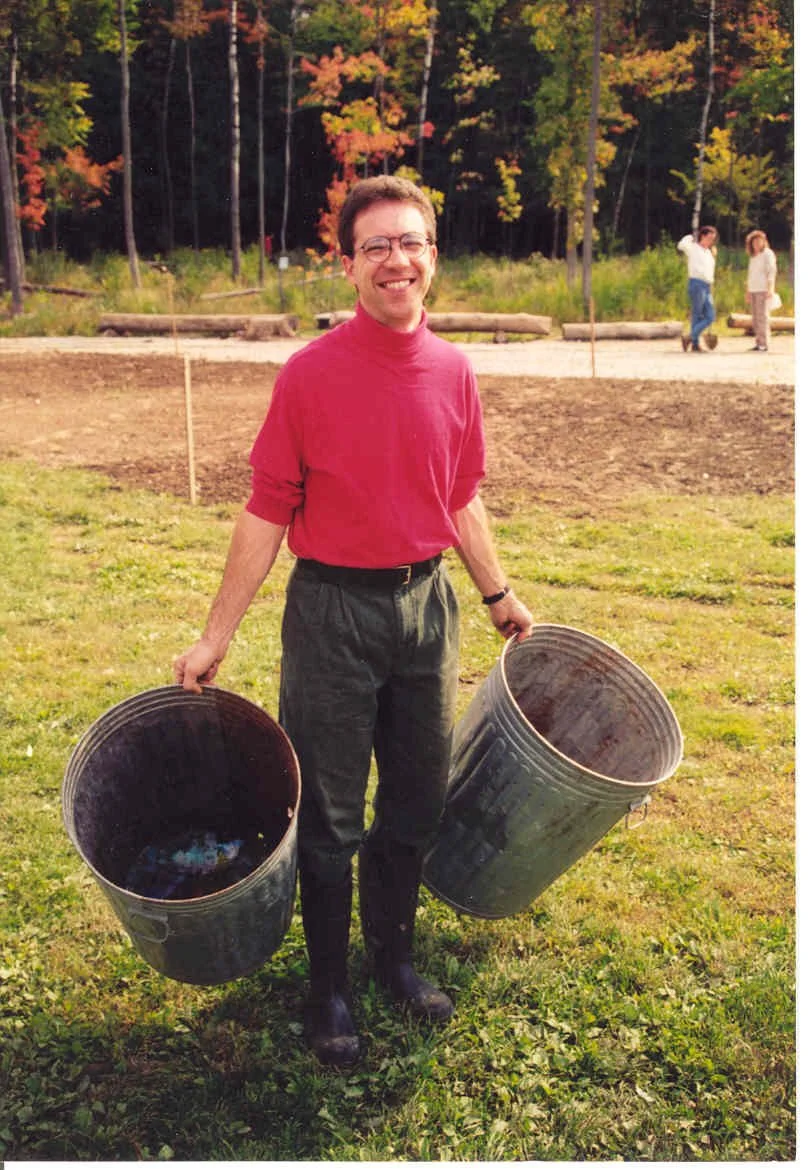 A man smiling, wearing glasses and a red turtleneck, standing outdoors on grass, holding two black trash cans in each hand, with a wooded area with colorful fall foliage in the background.