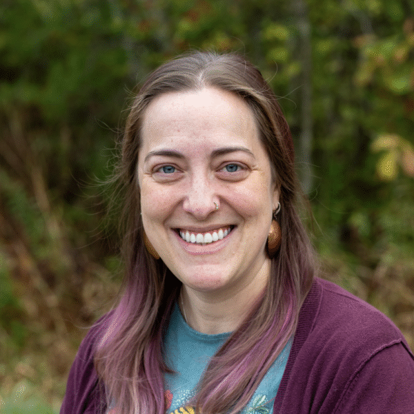 A woman with long, wavy hair dyed purple at the ends, smiling outdoors with green foliage in the background.