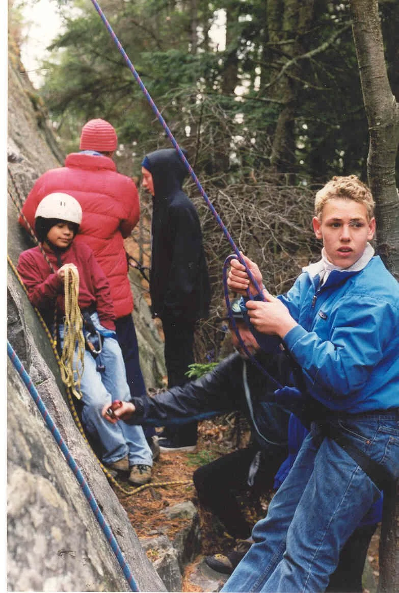 Group of children rock climbing outdoors in a forested area, some wearing helmets and jackets, with climbing ropes.