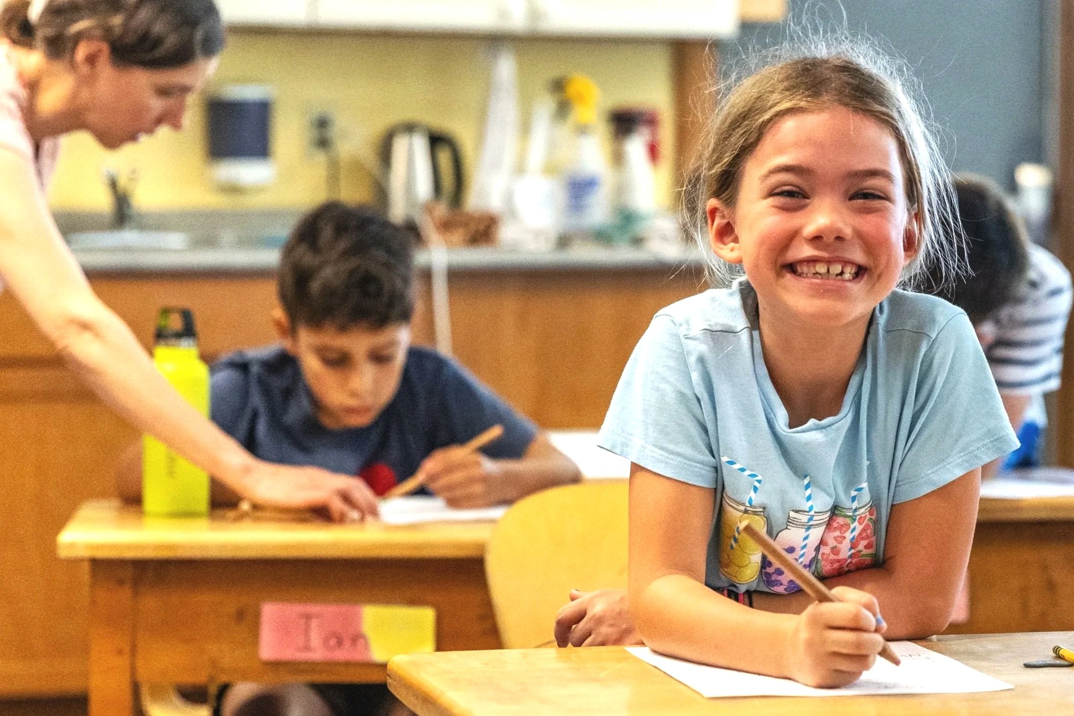 A young girl sits at a desk with a pencil in her hand smiling widely. A teacher and student are working together in the background.