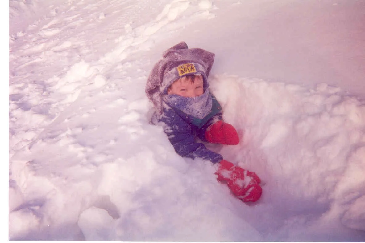 Young child playing in deep snow, wearing red gloves, a winter hat, and a face covering