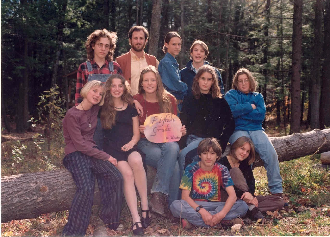 Group of nine children and one adult outdoors in a wooded area, with one girl holding a sign that reads "Eighth Grade Mr. Warren."