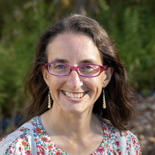 A smiling woman with wavy brown hair and pink glasses, wearing a colorful floral top and dangling earrings, outdoors in a natural setting.