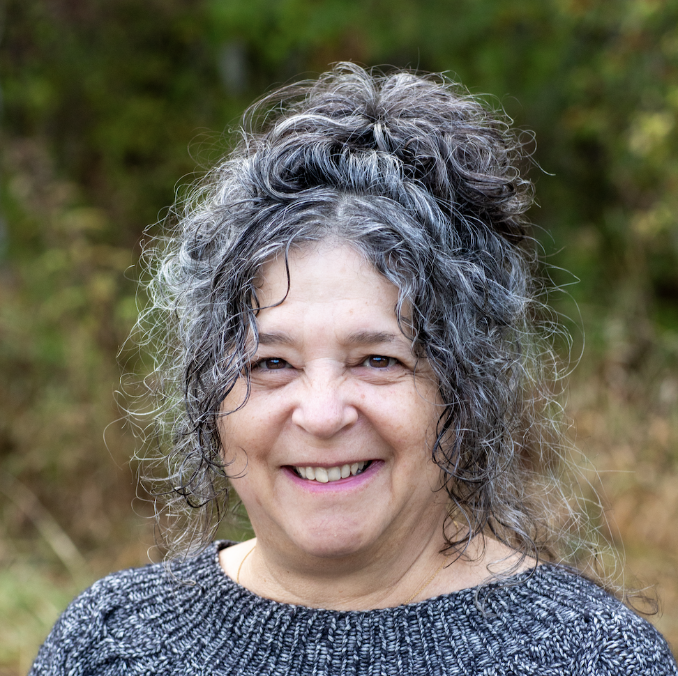 A woman with curly gray hair smiling outdoors, wearing a gray sweater, with a blurred forest background.