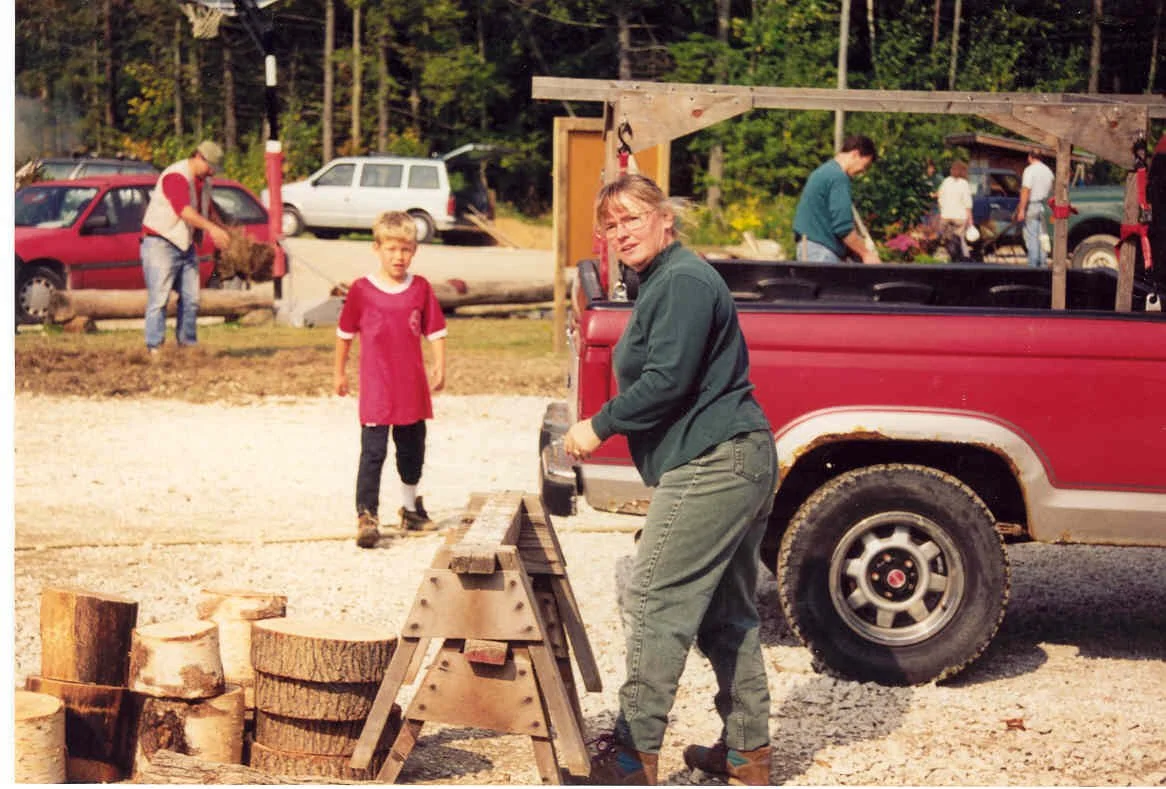 People outdoors at a rustic setting with logs, trees, and vehicles, including a woman sawing wood, a young boy in a red shirt, and several others in the background.