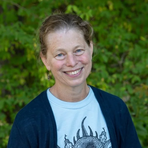 Woman with light brown hair and blue eyes smiling outdoors, wearing a white t-shirt and navy cardigan, with green foliage in the background.