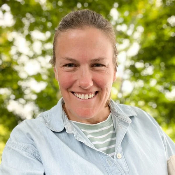 Smiling woman with light brown hair tied back, wearing a light denim shirt over a striped t-shirt, outdoors with blurred green trees in the background.