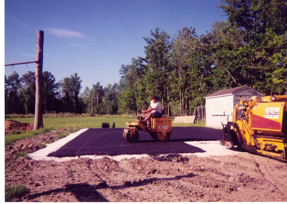 Person operating a roller compactor on a newly paved asphalt surface in a rural outdoor setting with trees and a shed.