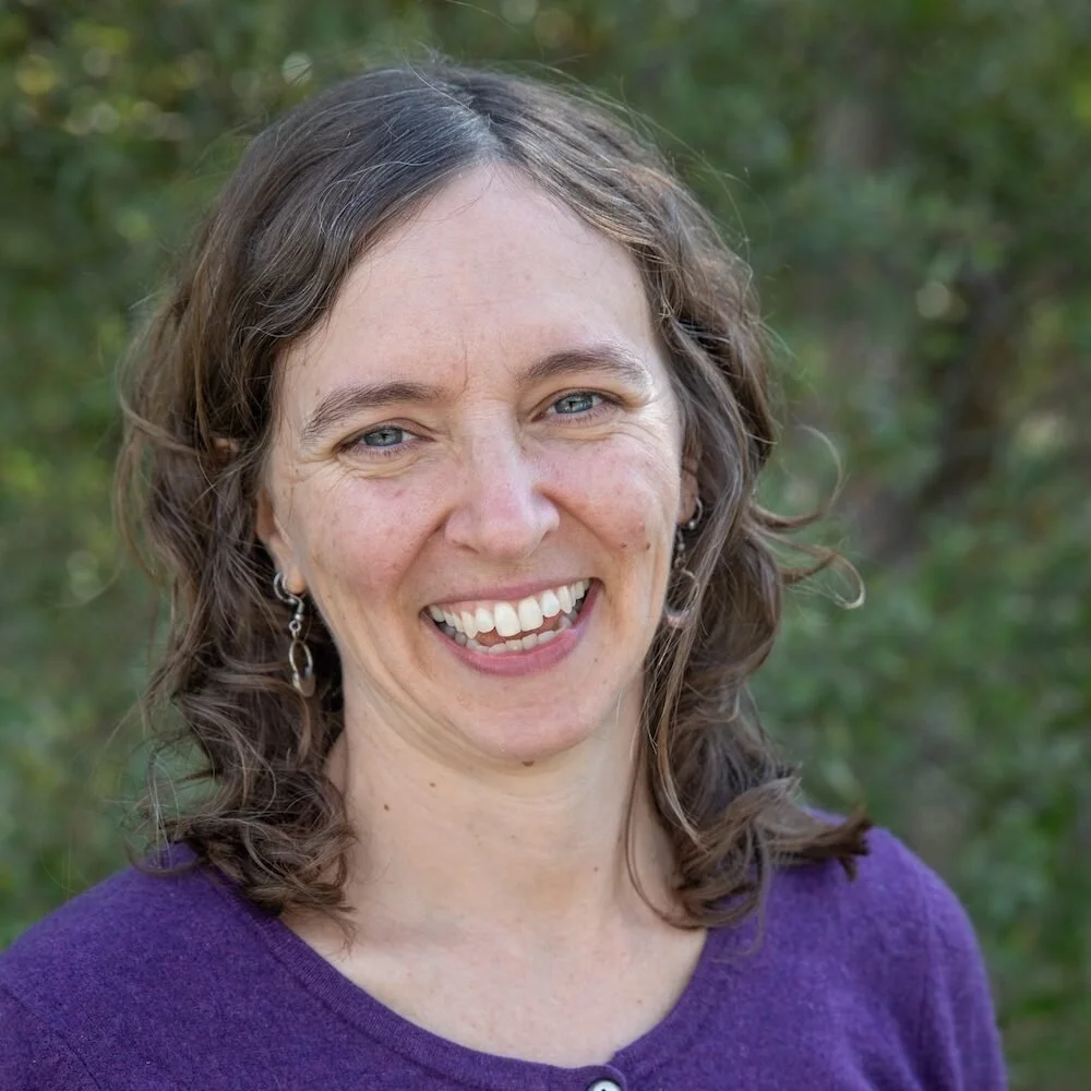 A woman with curly brown hair and blue eyes smiling outdoors, wearing a purple top and earrings.
