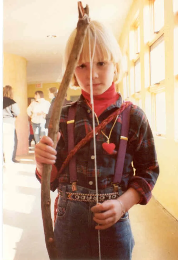 A young girl with blonde hair wearing a plaid shirt, red turtleneck, and suspenders, holding a stick in a brightly lit indoor space with windows and other people in the background.