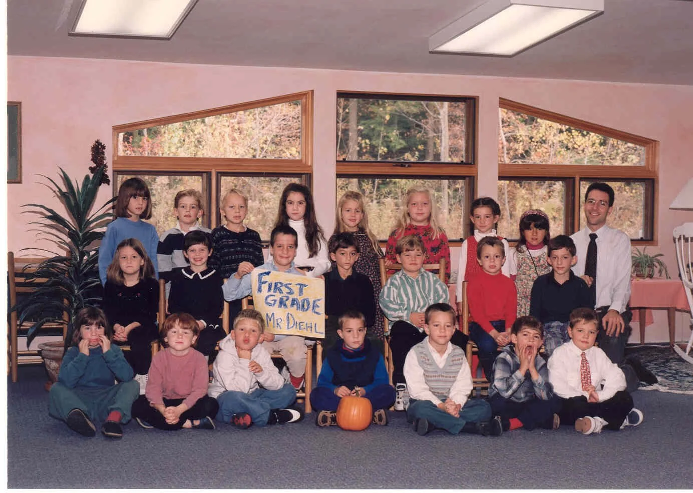 A classroom group photo of first-grade students and their teacher in a school, with a sign reading 'First Grade Mr Diehl' held by a student in the front row. The children are sitting and standing indoors, with large windows showing trees outside.