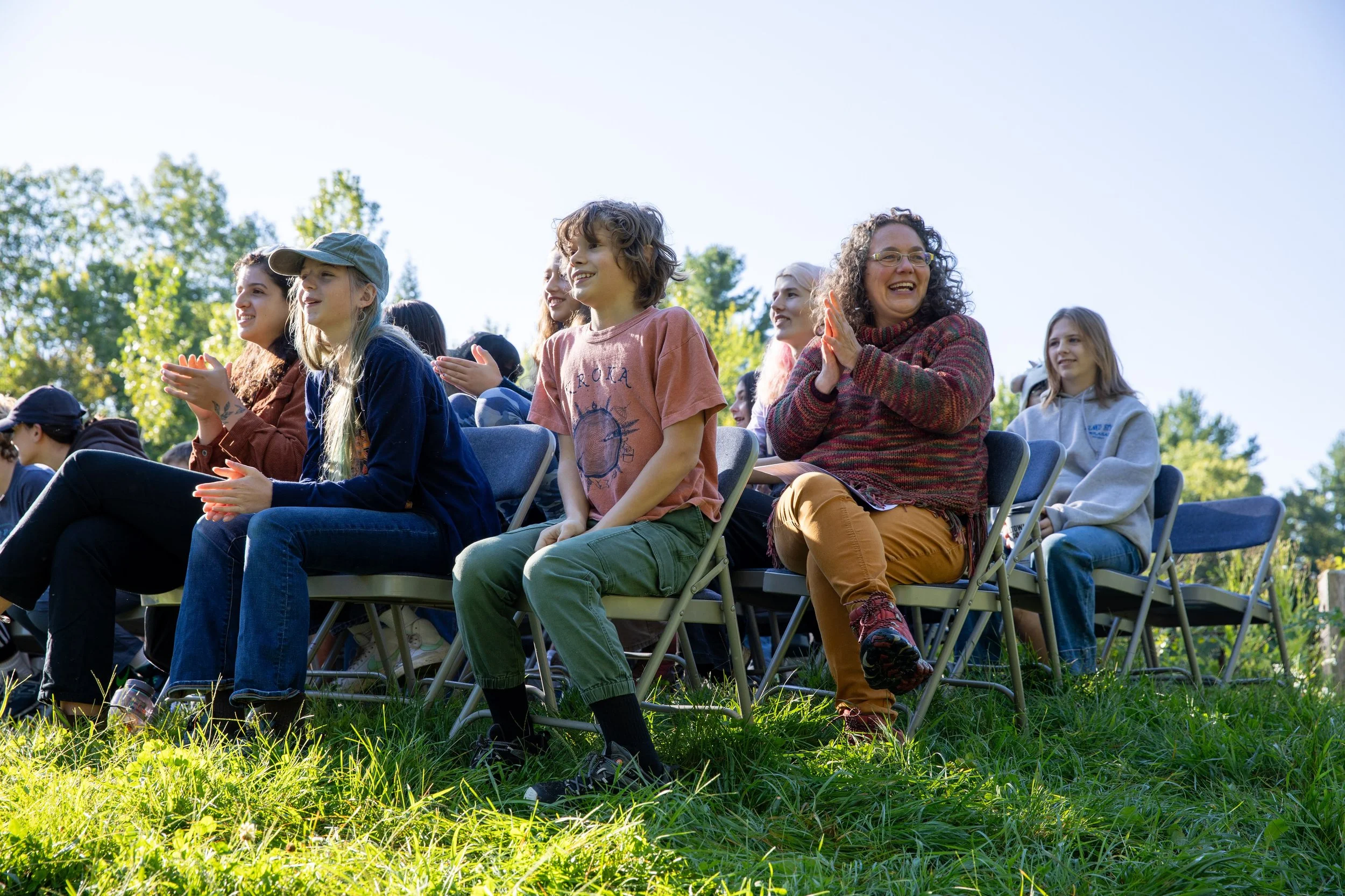 Teacher sits with students outdoors watching a performance and clapping