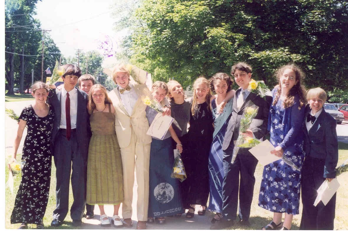 A group of people celebrating, some holding flowers and certificates, outdoors on a sunny day with trees in the background.