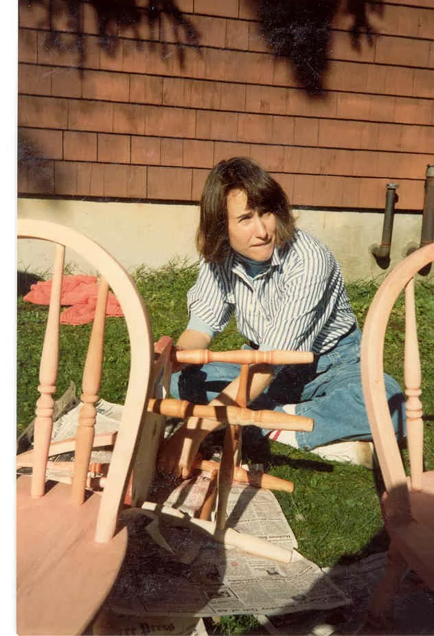 A woman with shoulder-length dark hair and a striped shirt is kneeling on the grass outside, dismantling wooden chairs. There are overturned chairs, newspapers on the ground, and a red cloth or towel nearby. The background features a wooden fence and