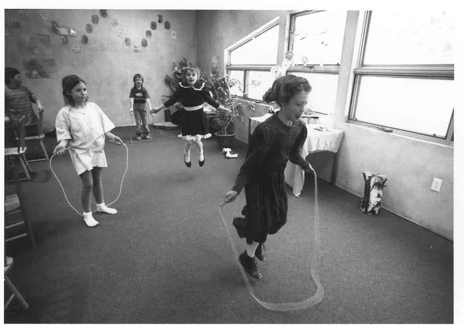 Children playing jump rope indoors in a bright room with large windows.