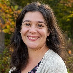 A woman with long dark hair smiling outdoors in front of trees with fall foliage.