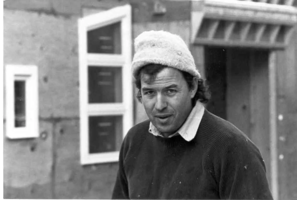 Black and white photo of a man wearing a knitted hat and a dark sweater, standing outside a building with a window.