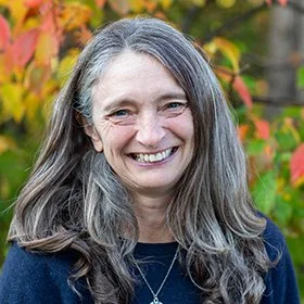Smiling woman with long gray hair wearing a dark top outdoors with colorful fall foliage in the background.