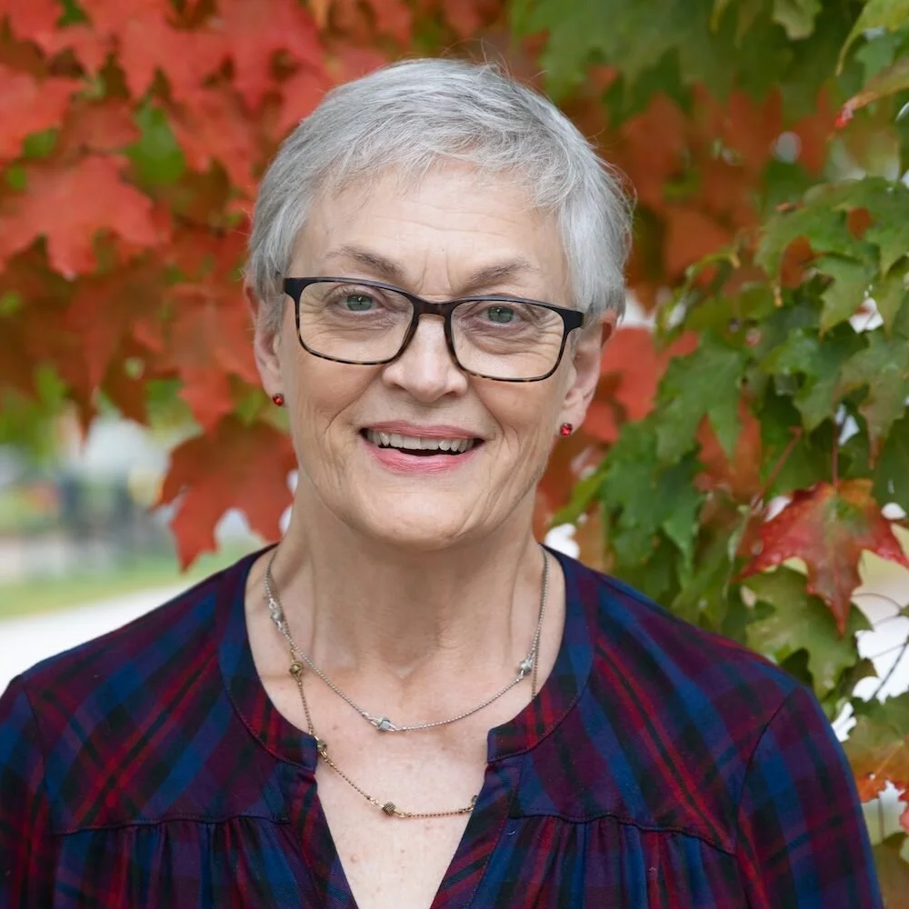 An elderly woman with short gray hair, wearing glasses, small earrings, and a necklace, smiling outdoors with red and green autumn leaves in the background.