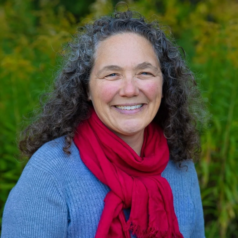 A middle-aged woman with curly, shoulder-length dark gray hair, smiling outdoors in front of green foliage, wearing a blue sweater and a red scarf.