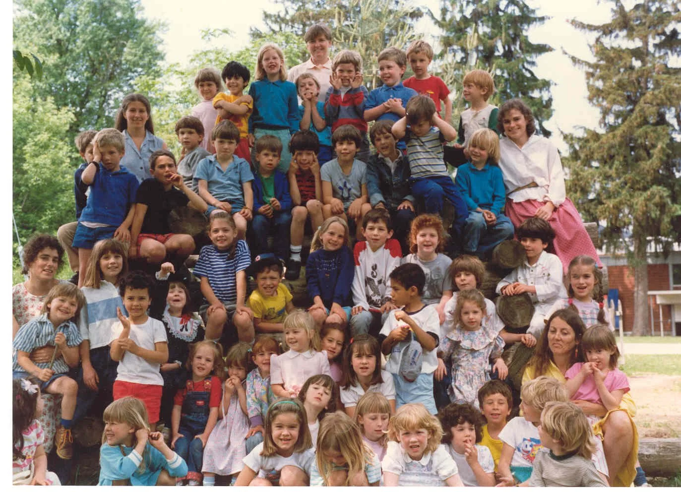 Group of school children and teachers outdoors on a large wooden structure, smiling and looking at the camera, with trees and a school building in the background.