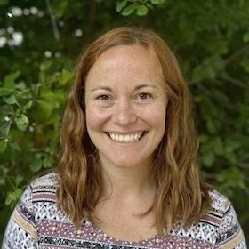 A woman with shoulder-length reddish-brown hair smiling outdoors with green foliage in the background.