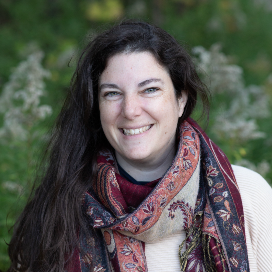 A woman with long dark hair and light skin smiling outdoors, wearing a patterned scarf.