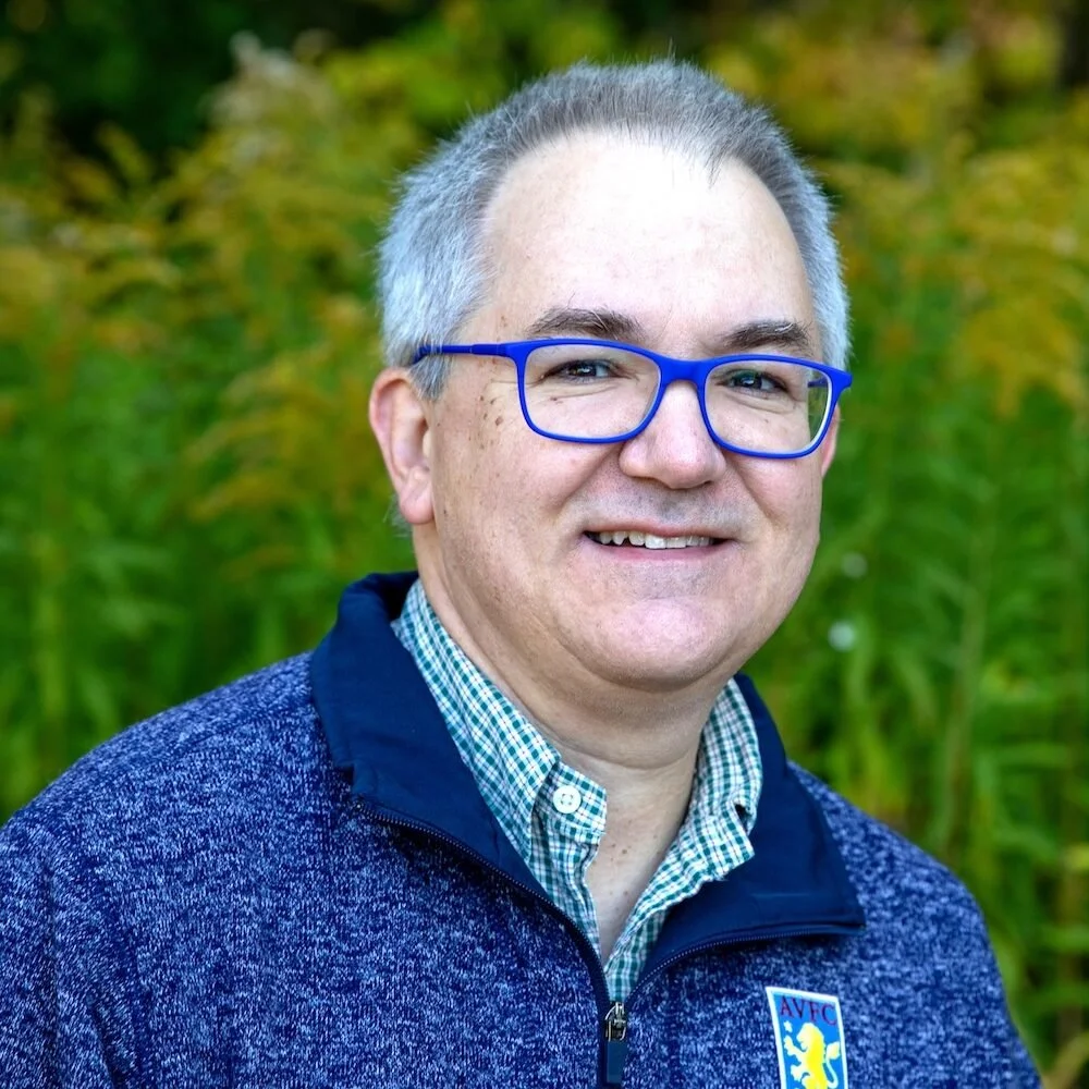 A man with short gray hair and blue glasses smiling outdoors in front of green foliage. He is wearing a checkered shirt and a dark blue zip-up jacket with a logo on it.