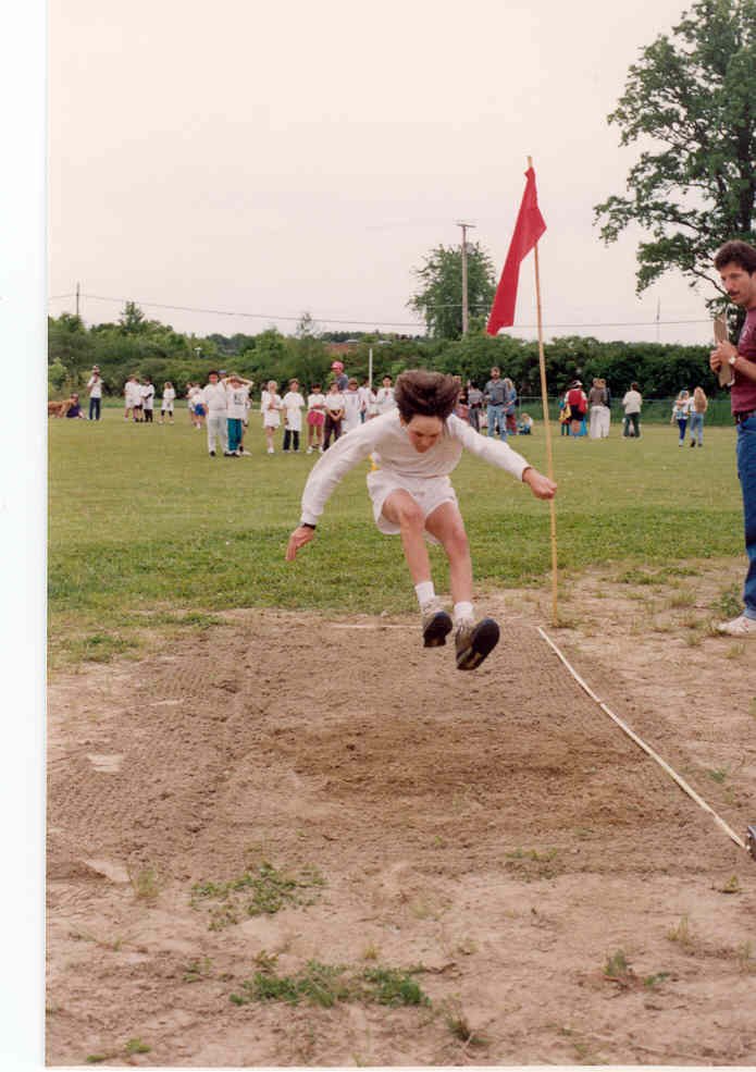 Young girl in white shorts and shirt jumping into a sandpit for a long jump event at an outdoor sports day.
