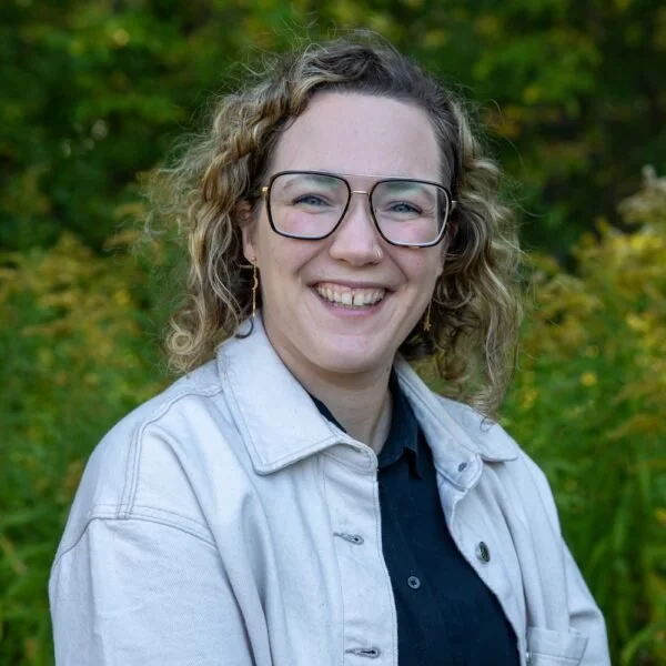 A woman with curly blonde hair, glasses, and earrings, smiling outdoors with green trees and foliage in the background.