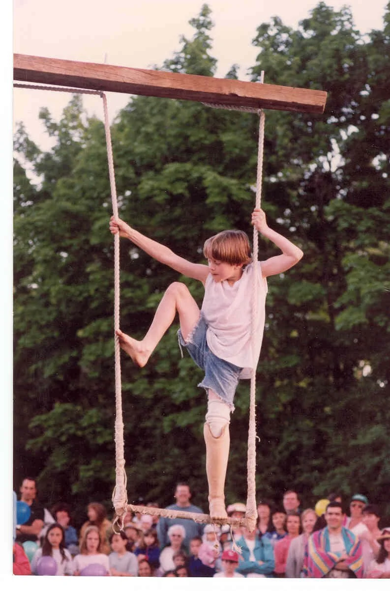 A boy with a mop of brown hair swinging on a wooden swing with rope seats in front of a crowd of onlookers and trees.