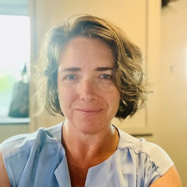 A smiling woman with short, wavy brown hair in a light blue top sitting indoors near a window.