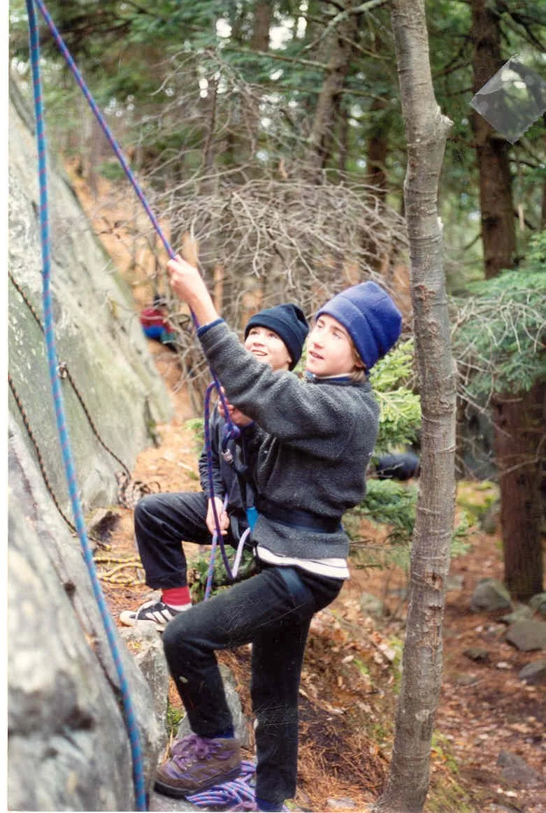 Two children, dressed in winter outdoor clothing, rock climb on a forested trail, using ropes for safety.