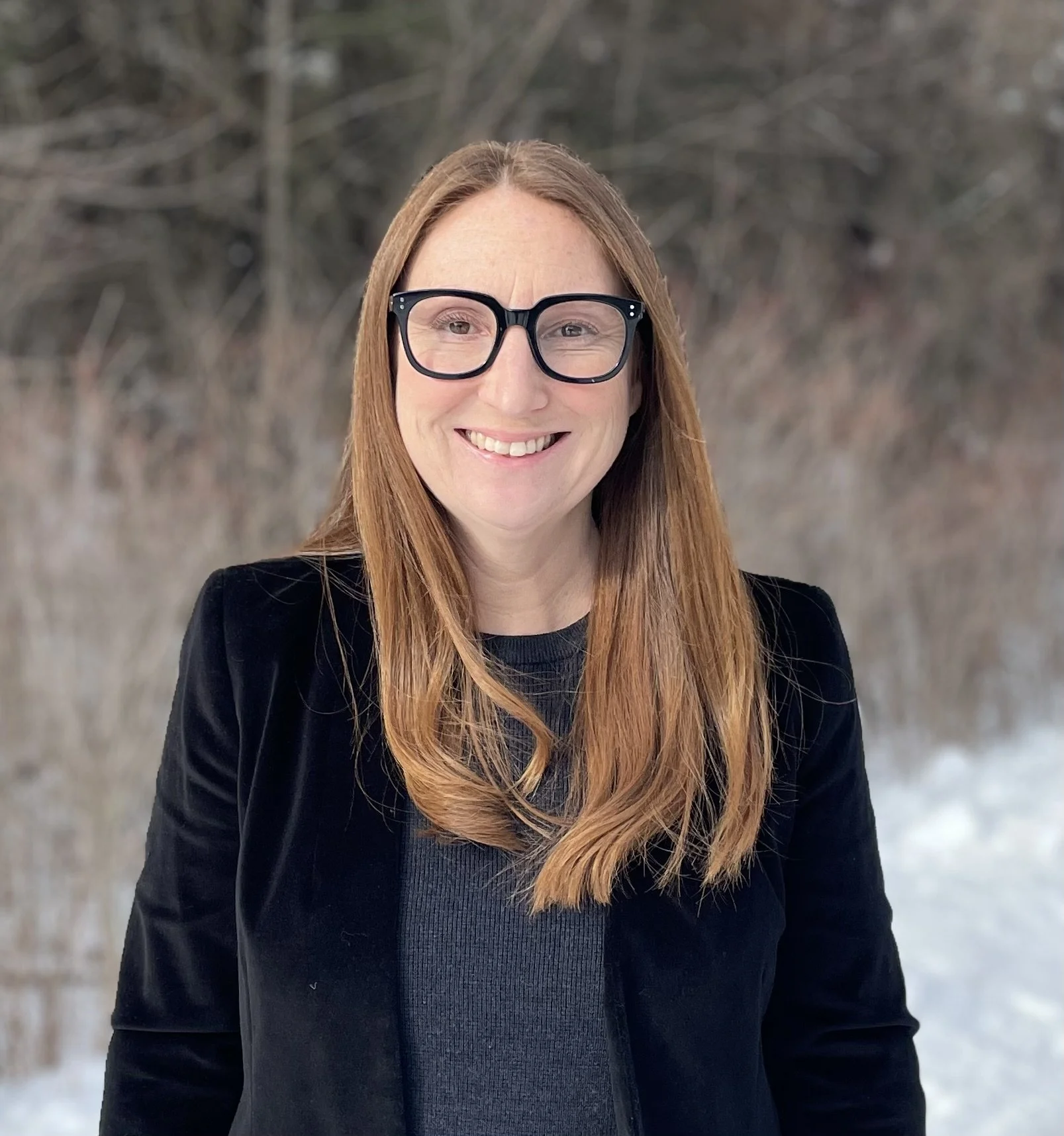 A woman with long red hair wearing glasses and a black blazer, smiling outdoors. The background features blurry trees and snow.