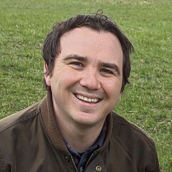A man with dark hair and a big smile, wearing a brown jacket, sitting outdoors on green grass.