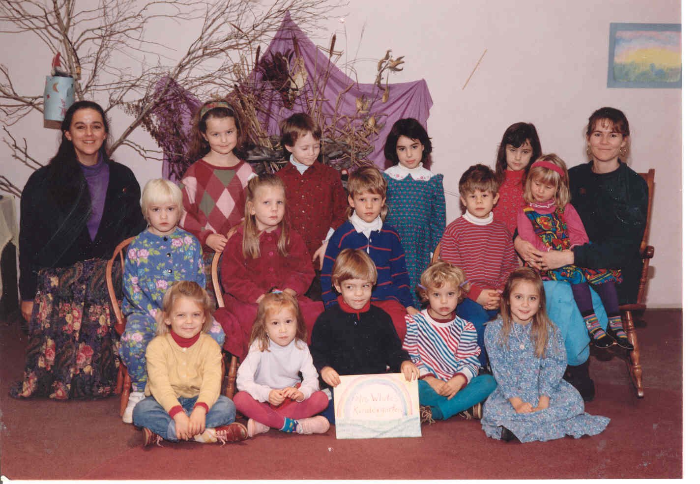 Group of young children and two adult women in a classroom, posing for a photo with a decorative backdrop and a rainbow painting on the wall.