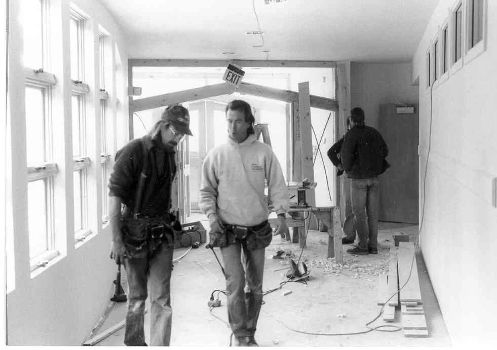 Three construction workers in a room under renovation, with one in the foreground and two in the background, surrounded by construction equipment and debris.