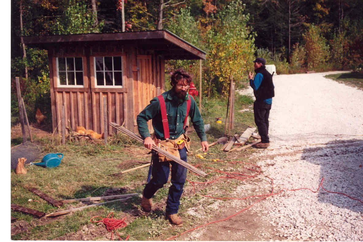 A man with a beard and glasses, carrying wood, stands on a gravel path near a small wooden shed, with another man standing further back making a gesture with his hand. There are trees with autumn foliage in the background, and various tools and mater