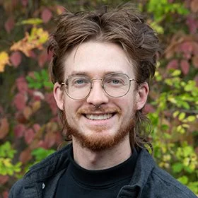 A person with glasses, a beard, and long hair smiling outdoors with autumn-colored foliage in the background.