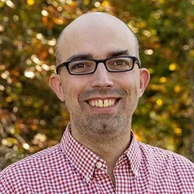A smiling man with glasses, wearing a red and white checkered shirt, standing outdoors with autumn foliage in the background.